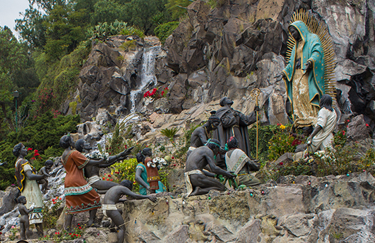 Photo of Our Lady of Guadalupe on top of a hill as her followers are looking up at her. Photo Courtesy of capitalbus.com.mx